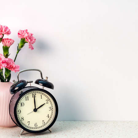 A square photo of terrazzo desk on white empty copy space wall with black alarm clock and pink carnations in a vaseの写真素材