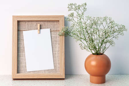 Desk at an empty white wall with a wooden frame with copy space and a vase with white fine flowersの写真素材