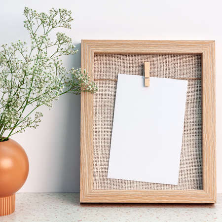 A wooden natural frame and an orange vase with an original shape with white flowers against a white...