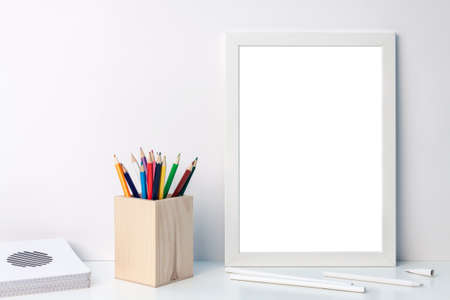 Photo of a white mockup frame with colored pencils in a wooden toolbox with notebooks on the deskの写真素材