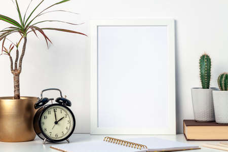 A white desk at home with plants in pots and a white mockup frame. Cactuses and yuka in a golden casing. Hipster decoration. A place for your text or graphics. Opened notebook, book and retro alarm clockの写真素材