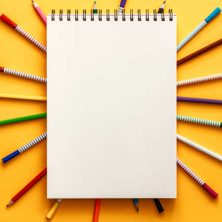 Yellow background with an empty sketchbook as a mockup. Colorful pencils and markers scattered around. Concept desk. Place for text. Top view, real photoの写真素材