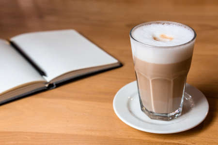 Wooden table with latte coffee and an open notebook for notesの写真素材