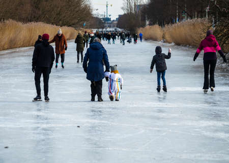 AMSTERDAM, THE NETHERLANDS, FEBRUARY 14,2021. Little toddler in a striped winter suit and woolen hat hold by the hand of a grandparent while walking on a frozen canal in Amsterdam, The Netherlands, surrounded by skating peopleのeditorial素材