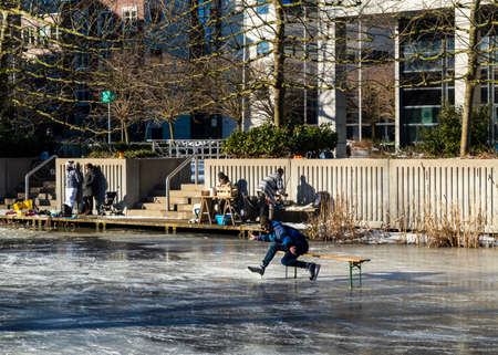 AMSTERDAM, THE NETHERLANDS, FEBRUARY 13,2021. Ice skater on a wooden bench on the frozen ditch before a building striking a funny pose and people in the background preparing warm drinksのeditorial素材