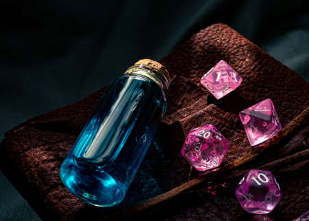 Close-up image of pink transparent role-playing gaming dice and a glass stopper bottle on top of a leather-bound book light by a ray of sunlightの写真素材