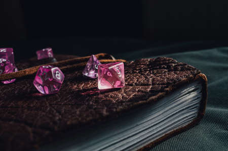 Close-up image of pink transparent role-playing gaming dice on top of a brown leather-bound book lit by a ray of sunlightの写真素材