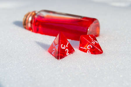 Close-up image of two red 4-sided role-playing game dice in the snow. In the background a glass stopper bottle with red liquidの写真素材
