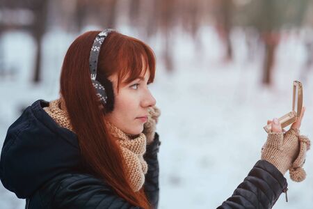 Winter. Woman with red hair looking in the mirror on the street. Female wearing ear muffs does make-upの写真素材