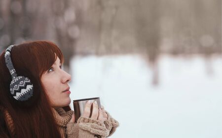 Winter. Woman with red hair wearing ear muffs. Girl drinking hot tea or coffee  iron insulated cupの写真素材