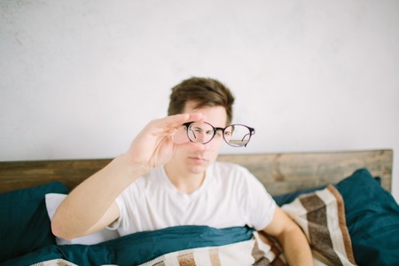 Closeup portrait of young man with glasses. He has eyesight problems and is squinting his eyes a little bit. Handsome guy is holding his eyeglasses right in front of camera with one handの写真素材