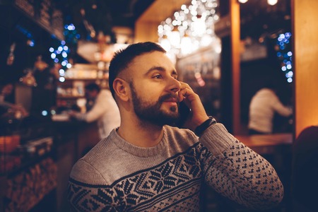 Side view of young bearded man,dressed incasual wear,sitting at round wooden table in cafe with modern interior and is holding smartphone.の写真素材
