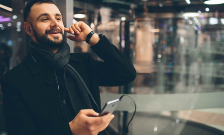 Side view of young bearded man,dressed incasual wear, he is standing on the street and using a smartphone and listening to musicの写真素材