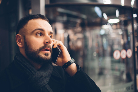Side view of young bearded man,dressed incasual wear, he is standing on the street and using a smartphoneの写真素材