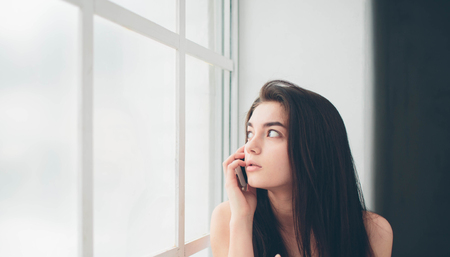 A young woman with black hair in a white T-shirt at a window flooded with sunlight uses a smartphone, communicates and searches for information on the Internet or social networksの写真素材