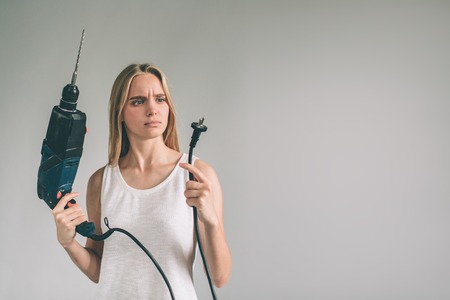 Funny portrait upset craftswoman. Blonde woman is wearing shirt isolated on white. Girl does not know how to use a drillの写真素材