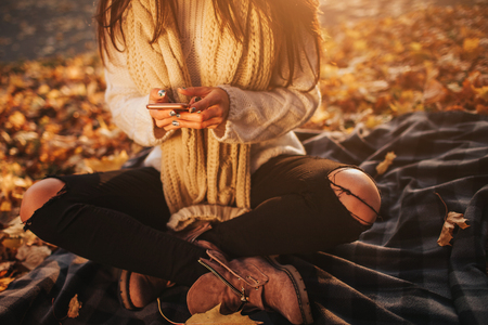 Woman using smartphone in fall. Autumn girl having smart phone conversation in sun flare foliage. Portrait of Caucasian model in forest in fall colorsの写真素材