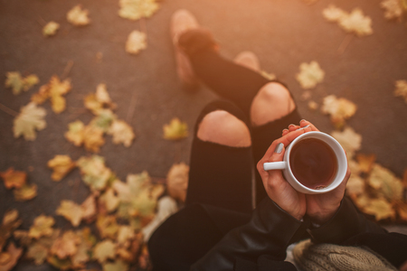 A young girl holding a cup of hot drink and smiling at the background of a autumn forest. Attractive young woman in autumn outdoor. Female model drinking hot tea in the autumn forest, close upの写真素材