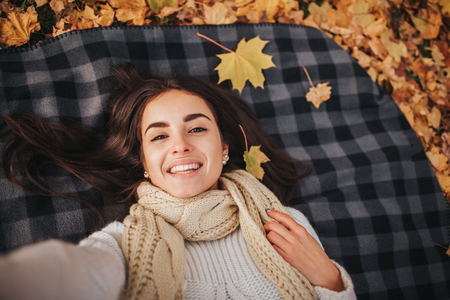 season, technology and people concept - beautiful young woman lying on ground and autumn leaves and taking selfie with smartphoneの写真素材