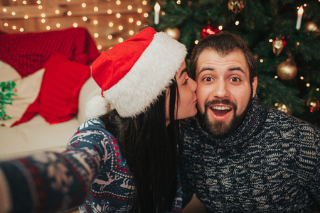 Merry Christmas and Happy New Year . Young couple celebrating holiday at home. Man and woman do selfie on smartphoneの写真素材