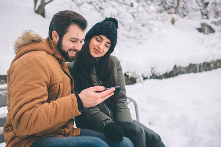 Happy Young Couple in Winter . Family Outdoors. man and woman looking upwards and laughing. Love, fun, season and people - walking in winter park. Sit on the bench. Holds a smartphone.の写真素材