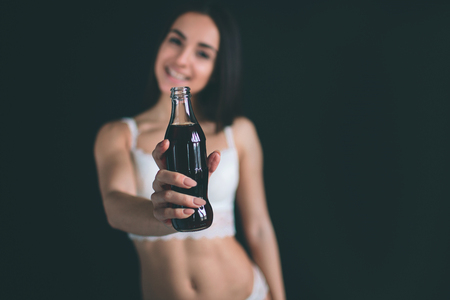 close up of happy woman drinking cola with straw. Young woman with long black hair stands isolated on black background. The girl has a sports figure, she is dressed in white underwear.の写真素材