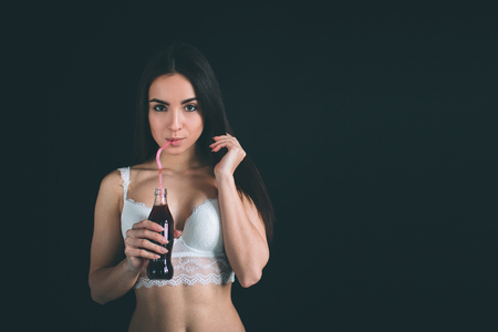 close up of happy woman drinking cola with straw. Young woman with long black hair stands isolated on black background. The girl has a sports figure, she is dressed in white underwear.の写真素材
