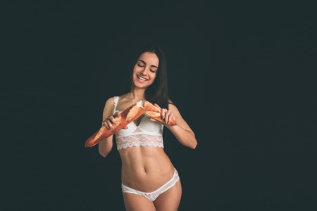 beautiful woman holding baguette. Vegetarian girl on a diet. Young woman have long black hair isolated on black background. The girl has a sports figure, she dressed in white underwear.の写真素材