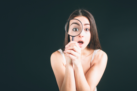 Shocked woman looking through a magnifying glass. Young woman with long black hair stands isolated on black background. The girl has a sports figure, she is dressed in white underwear.の写真素材
