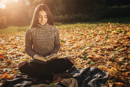 Beautiful young brunette sitting on a fallen autumn leaves in a park, reading a bookの写真素材