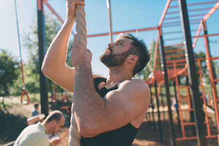 Bearded with black hair man climbing thick rope during fitness exercises in cross-fit training with....