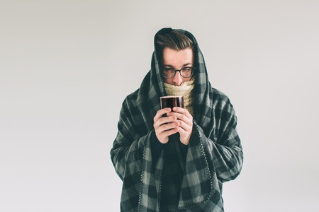 wrapped in the warm blanket or plaid, sick, young snotty man in glasses drinking warm healing tea.の写真素材