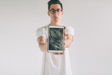 Nerd is wearing glasses. Student presenting a broken black tablet behind glass. upset man holds a out-of-use tablet or smartphone. Isolated on a light background. Broken screenの写真素材