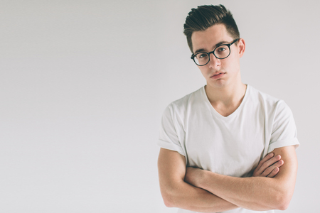 Portrait of young Nerd man wearing glasses and t-shirt standing with crossed arms and on white background while looking at the camera with disappointed, offended, and angry look on his face.の写真素材