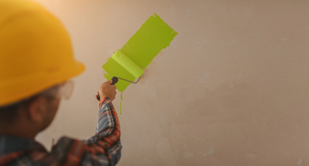 The builder works at the construction site and measures the ceiling. A worker in an orange helmet and a paint roller paints the wall.の写真素材