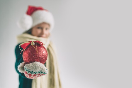 Big red Christmas ball in hands at the girl. The child is dressed in sweater, christmas hat and scarf studio shotの写真素材