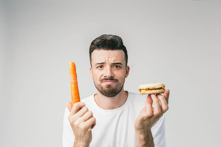 Close up of a bearded guy standing in the white room and holding a carrot in his right hand and a small burger in left hand.の写真素材