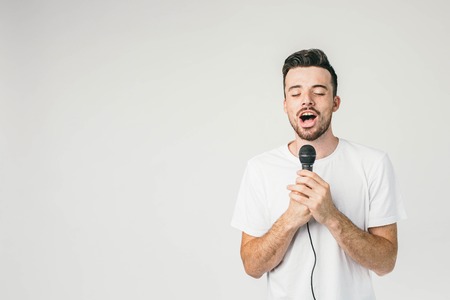 Brave guy standing at white wall and holding a microphone with his both hands. He is singing sensual and sad song with his eyes closed.の写真素材