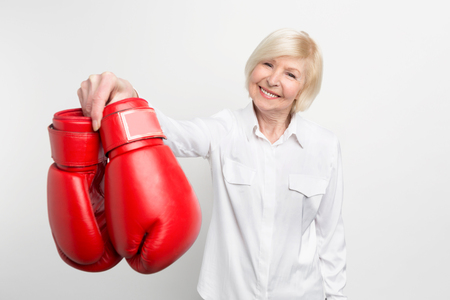 Joyful and nice old woman is holding boxing gloves in her right hand and smiling. She has what to do in her retirement. Isolated on white background.の写真素材
