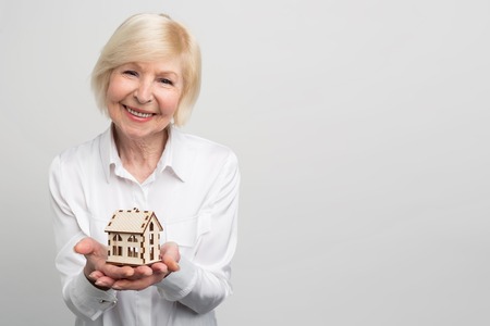 A picture of happy and smiley woman that has a small house-toy in her hand. She is not young right now and want to have some warranties for the future. Isolated on white background.の写真素材