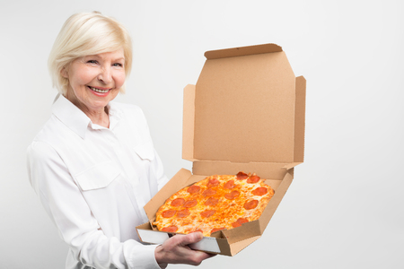 Cut view of grandma holding a big box of tasty pizza. She likes the junk food despice it is not healthy for people. Lady looks joyful and happy. Isolated on white background.の写真素材