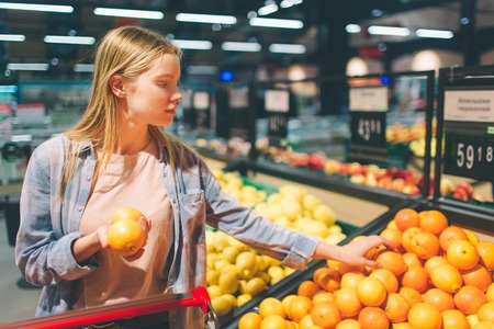 A picture of a girl standing near big basket of citrus. She has chosen one orage and trying to choose some more of it. There is a piece of gates on the photo. Cut view.の写真素材