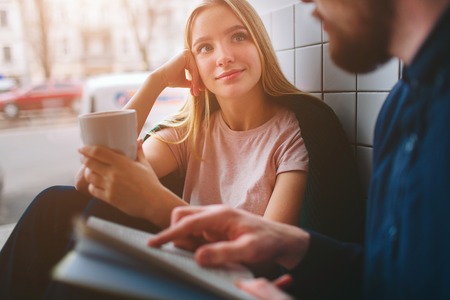 A girl sitting in cafe near big window and looking to her boyfriend. Se is listening to him very careful while he is reading a book for her. Close up. Cut view.の写真素材