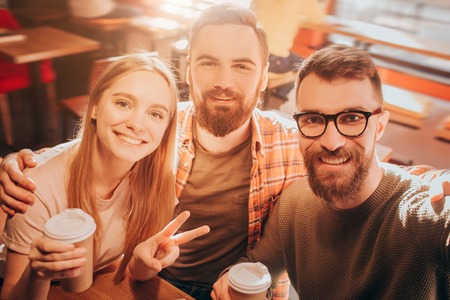 Nice selfie of two men and one woman sitting together very close and posing on camera. The girl shows the symbol of peace with her fingers. Cut view.の写真素材