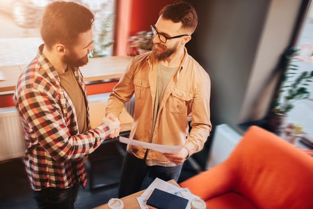 Two hipsters that wear casual clothes standing in a small cafe and shaking their hands.One of them is holding a document in his hand. Both of them are satisfied with their collaboration.の写真素材