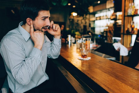 Dark-haired guy is sitting at the barmans stand and talking on the phone. He is closing his ear to hear what his friend is talking on the phone.の写真素材