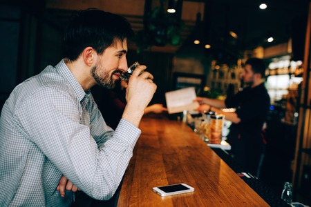 A guy sitting at the barmans stand and drinking some alcohol from the glass. He looks concentrated. He is smiling. Barman is giving a menu to a girl.の写真素材