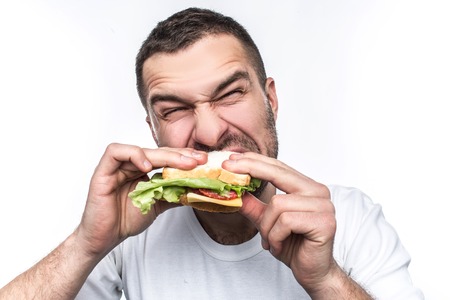 Funny and hungry guy is eating some fast food. He is hungry like a wolf. Man is biting sandwich very hard. Isolated on white background.の写真素材