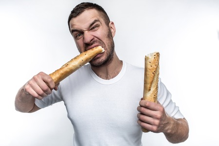 Very hungry man is starving. He broke a long piece of baguette into two pieces. He is eating one of this pieces. He looks brutal and weird. Isolated on white background.の写真素材