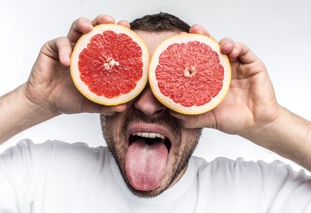 Funny and good picture of adult man putting two half pieces of grapefruit onto his eyes. Also he is showing his tongue to the camera. Isolated on white background.の写真素材